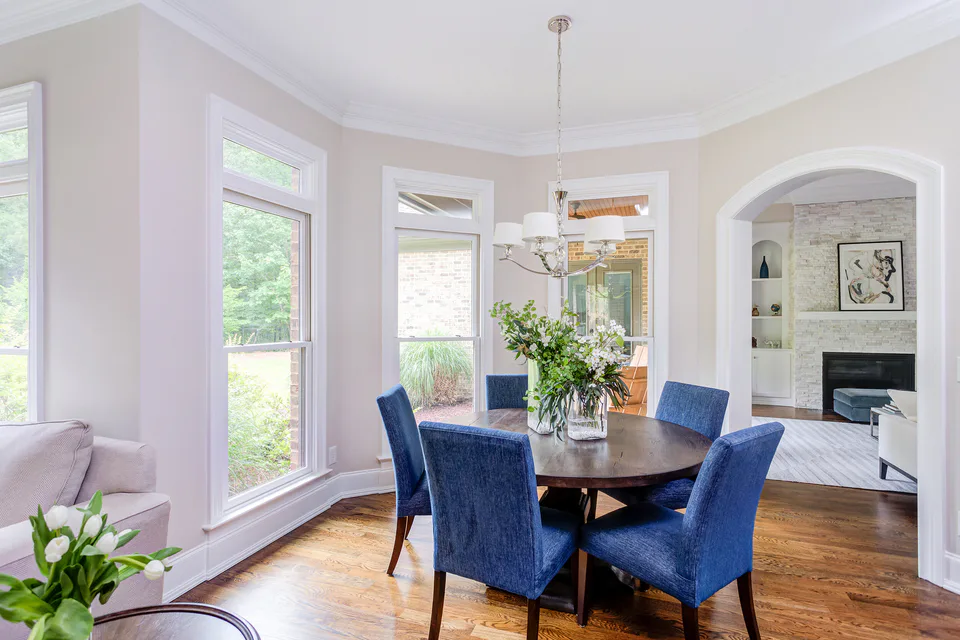 Breakfast room with a round dark wood table and blue chenille dining chairs beneath a nickel chandelier, bay windows and an arched opening to the living room at Durham Transitional, Durham, North Carolina, interior design by Alexandra Peck Design