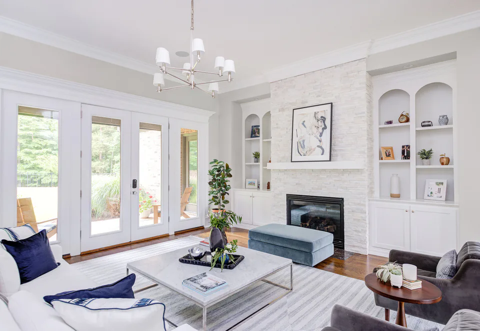 Living room with a white linen sofa, navy pillows, a marble coffee table, a blue velvet ottoman and a white stone fireplace between arched built-ins at Durham Transitional, Durham, North Carolina, interior design by Alexandra Peck Design