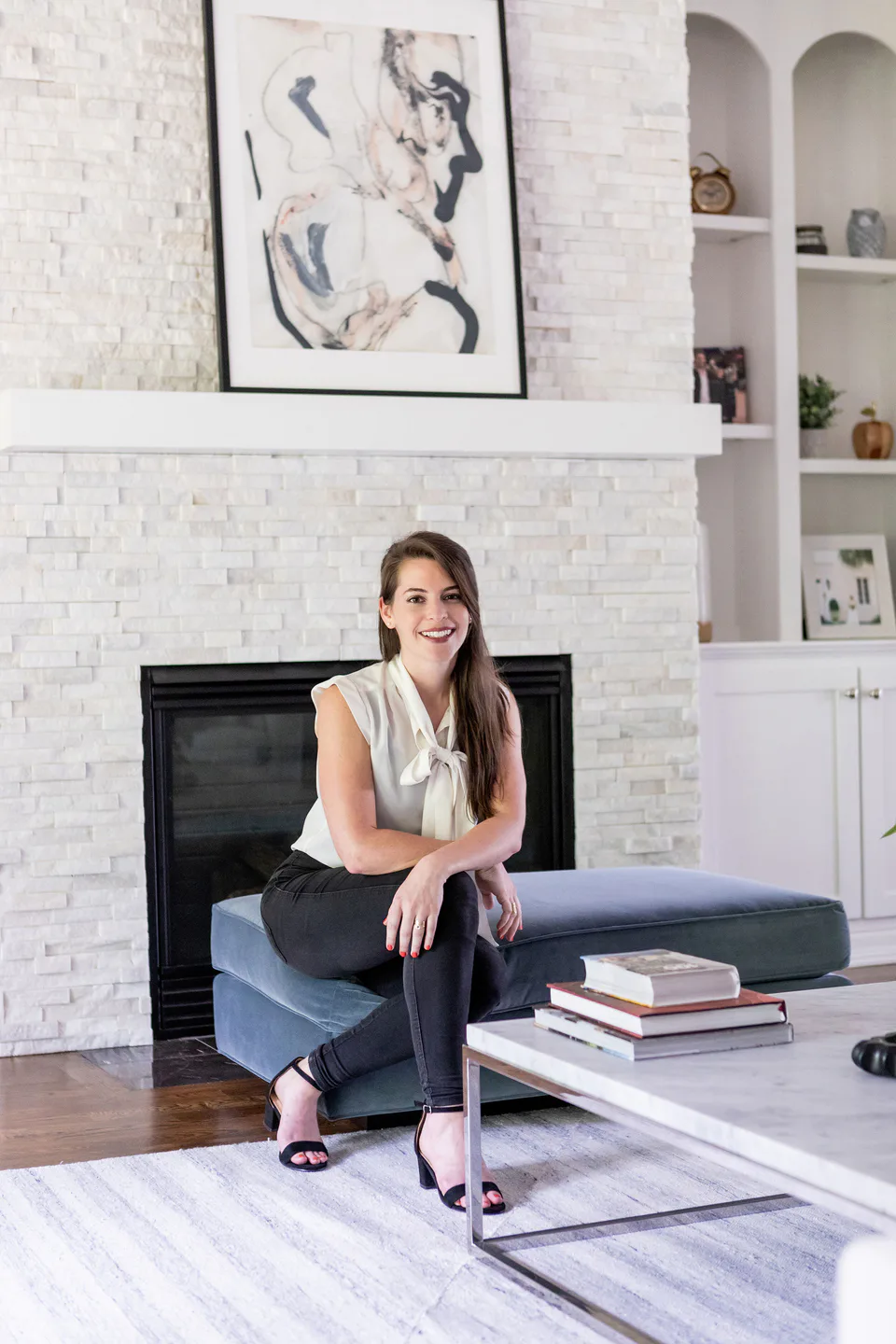 Alexandra Peck seated on the blue velvet ottoman by the white stone fireplace in the living room at Durham Transitional, Durham, North Carolina, interior design by Alexandra Peck Design