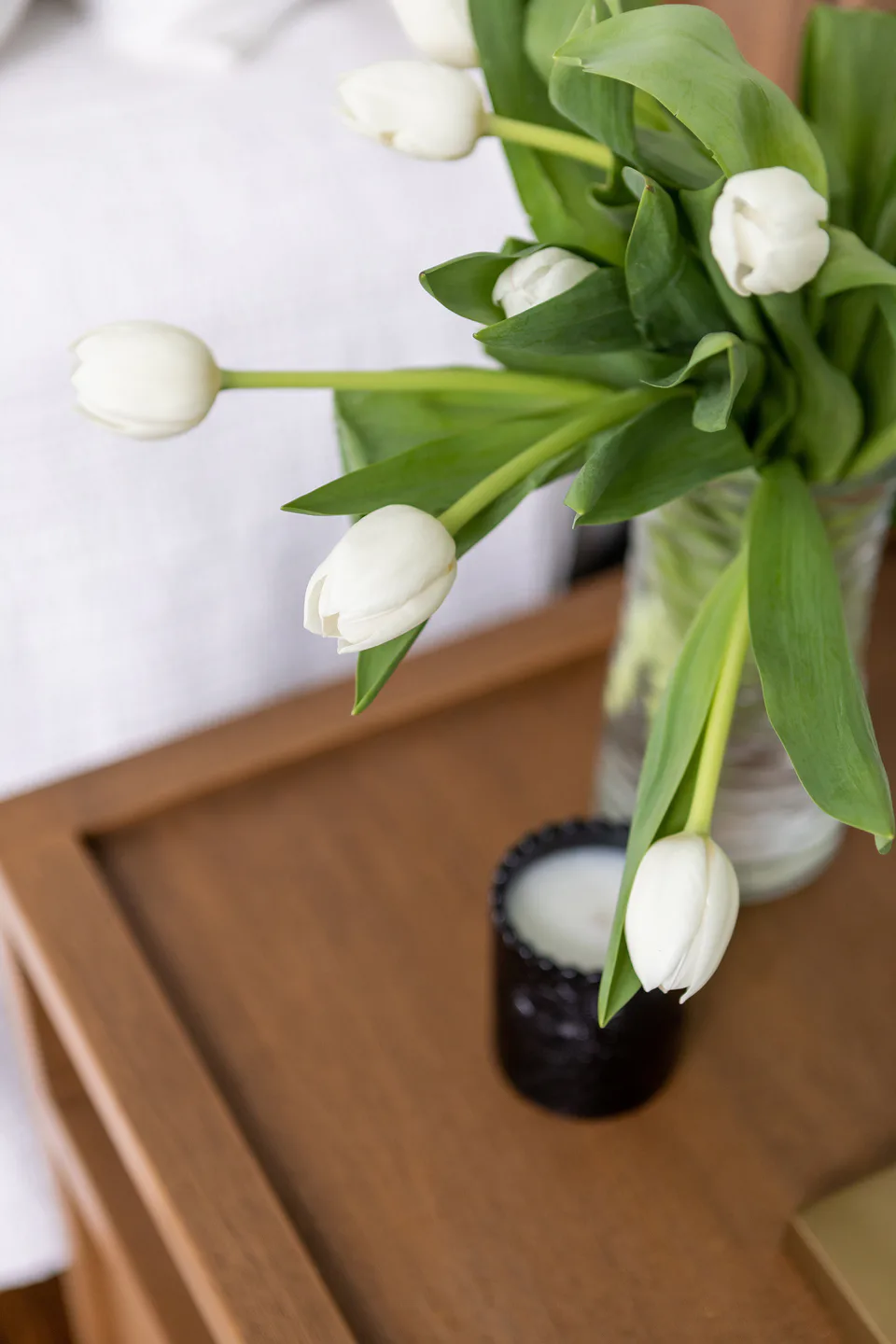 White tulips and a candle on the wood nightstand in the main bedroom at Durham Transitional, Durham, North Carolina, interior design by Alexandra Peck Design