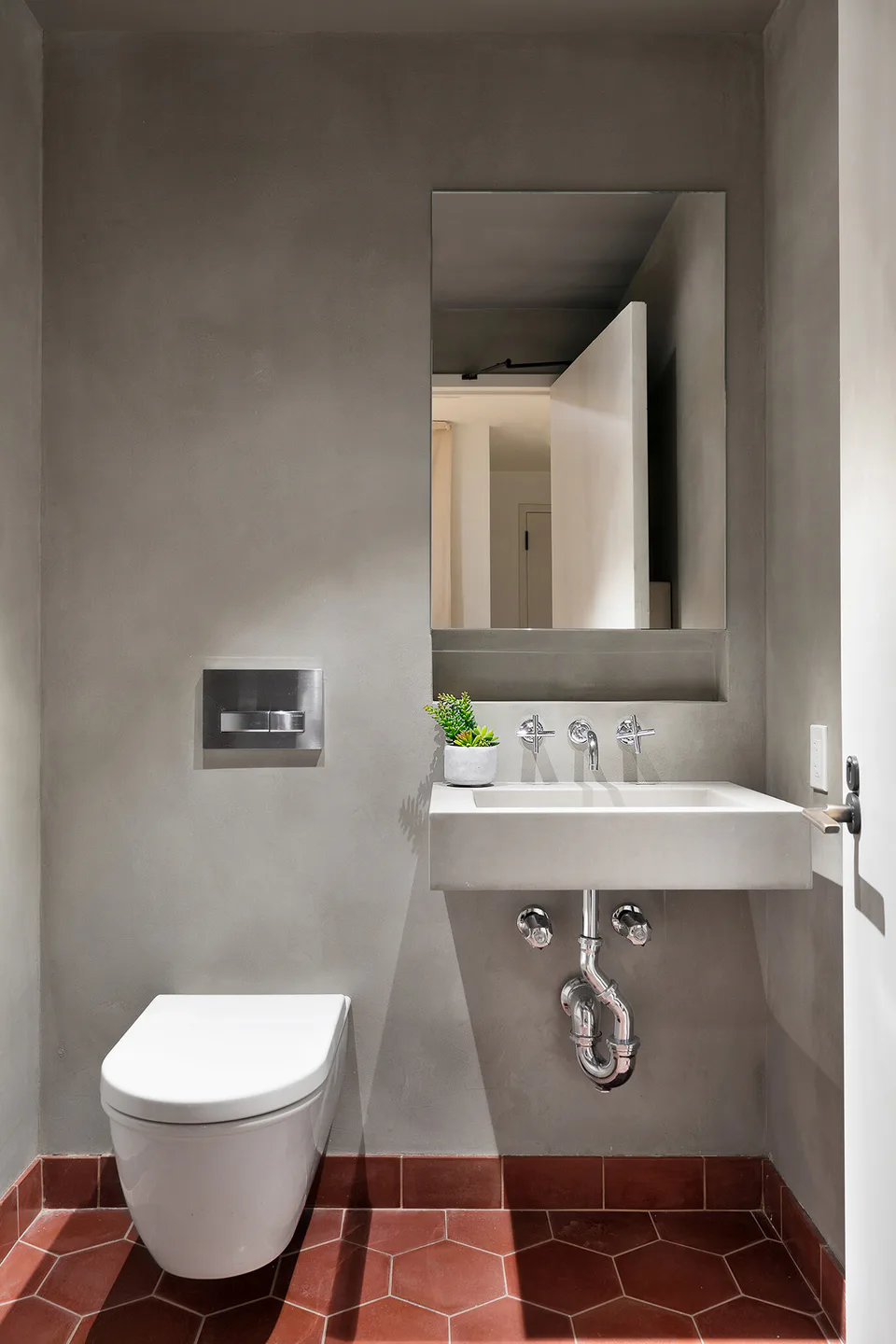 Restroom with gray plaster walls, a wall-hung sink and toilet and red hexagonal floor tiles at Flatiron Workplace, 3 East 28th Street, New York City, interior design by Alexandra Peck Design