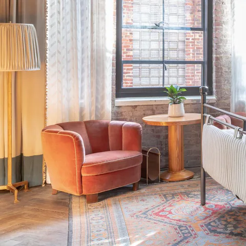 Detail: coral velvet armchair and a round side table beneath the factory window in a medium bedroom at Soho House Nashville, Nashville, Tennessee, interior design by Alexandra Peck Design
