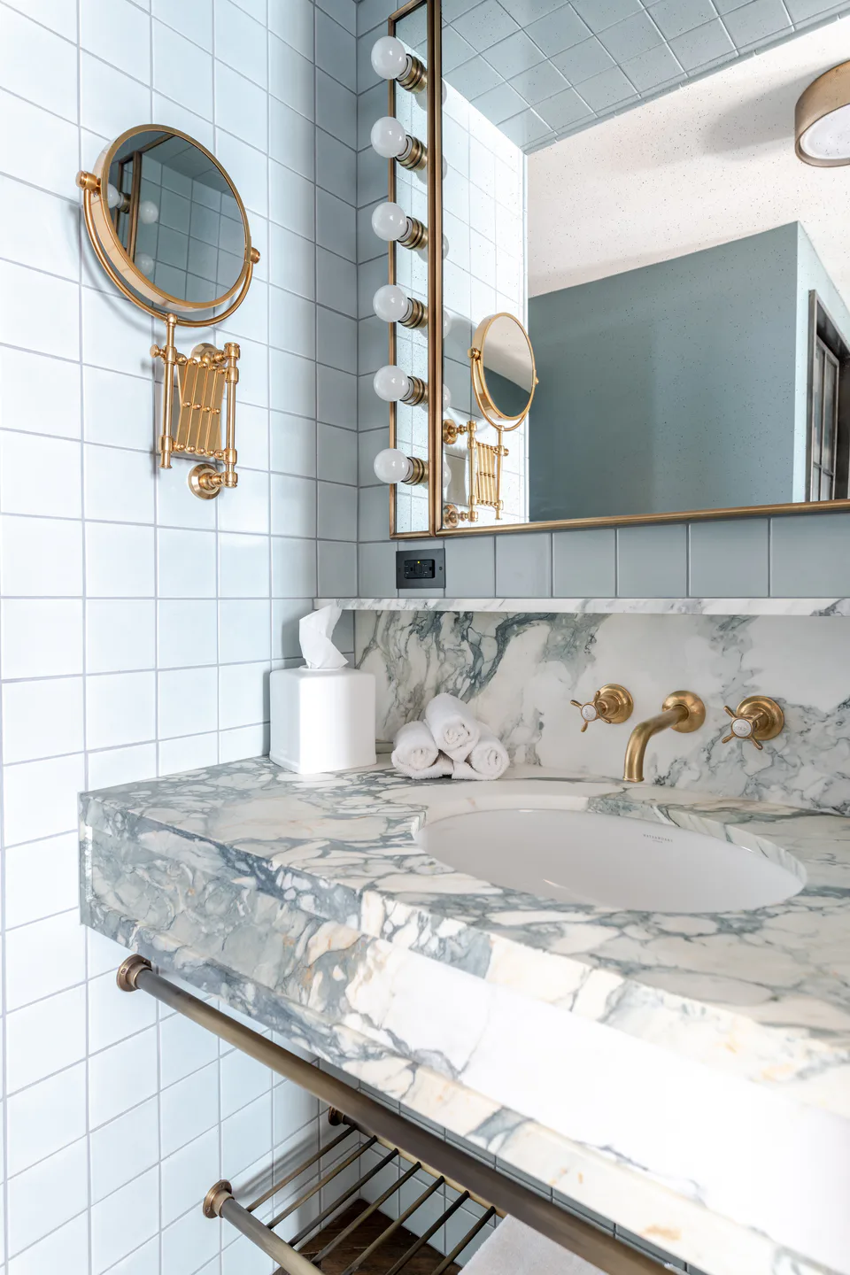 Marble basin with brass taps, a brass shaving mirror and pale blue tiles in a medium bathroom at Soho House Nashville, Nashville, Tennessee, interior design by Alexandra Peck Design