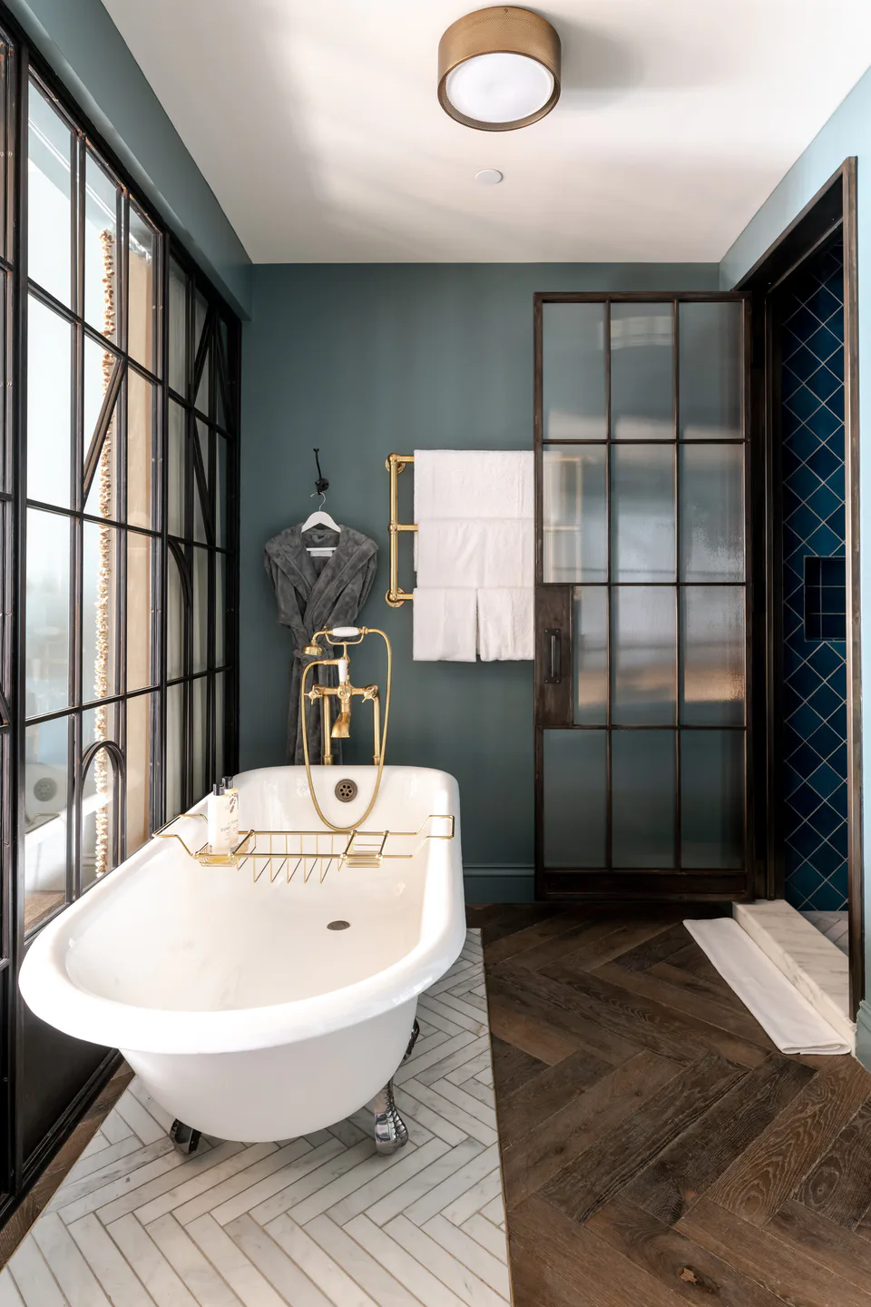 Clawfoot bathtub with brass fittings on a herringbone floor beside a steel-framed shower in a medium bathroom at Soho House Nashville, Nashville, Tennessee, interior design by Alexandra Peck Design