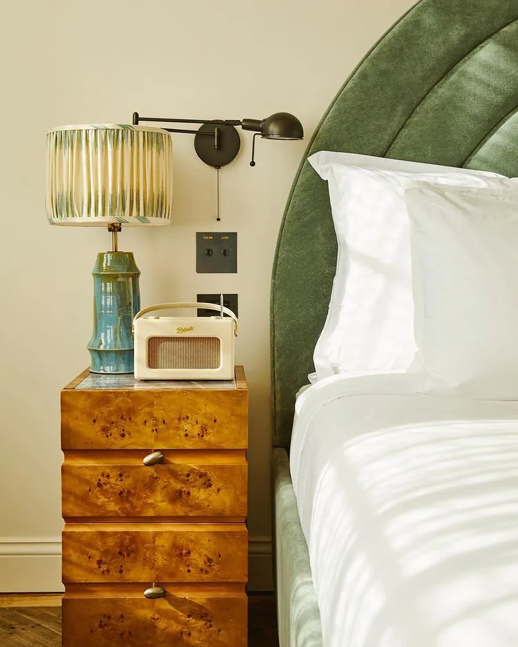 Burl bedside chest with a ceramic lamp and a radio beside the green velvet headboard in a small bedroom at Soho House Nashville, Nashville, Tennessee, interior design by Alexandra Peck Design