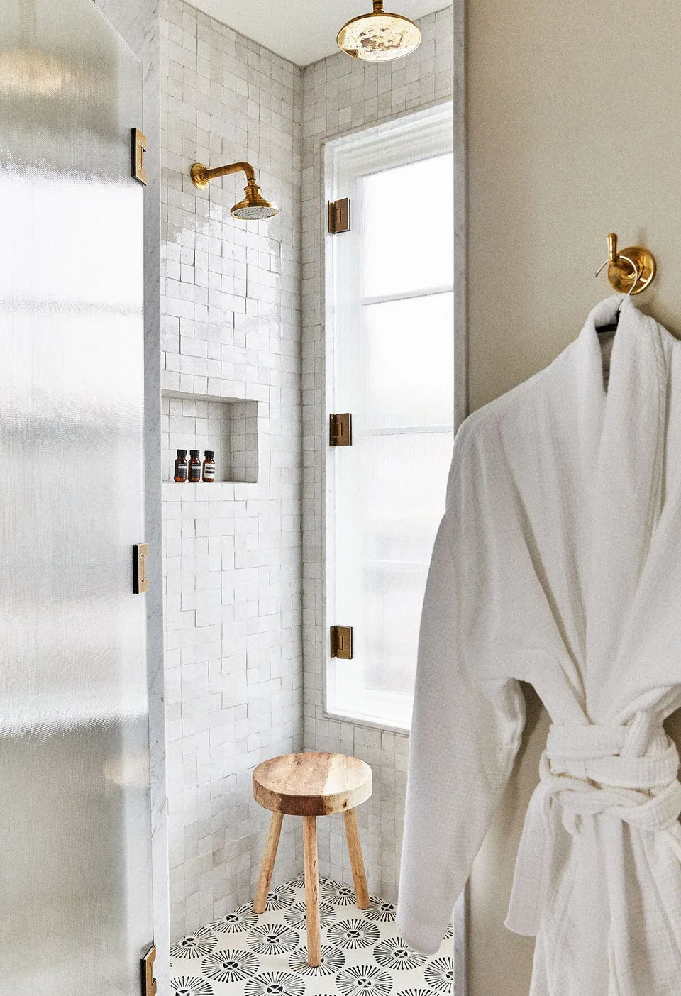 Bathroom with a white tiled shower, a wooden stool and a robe on a brass hook at The Pinch Hotel, Charleston, South Carolina, interior design by Alexandra Peck Design