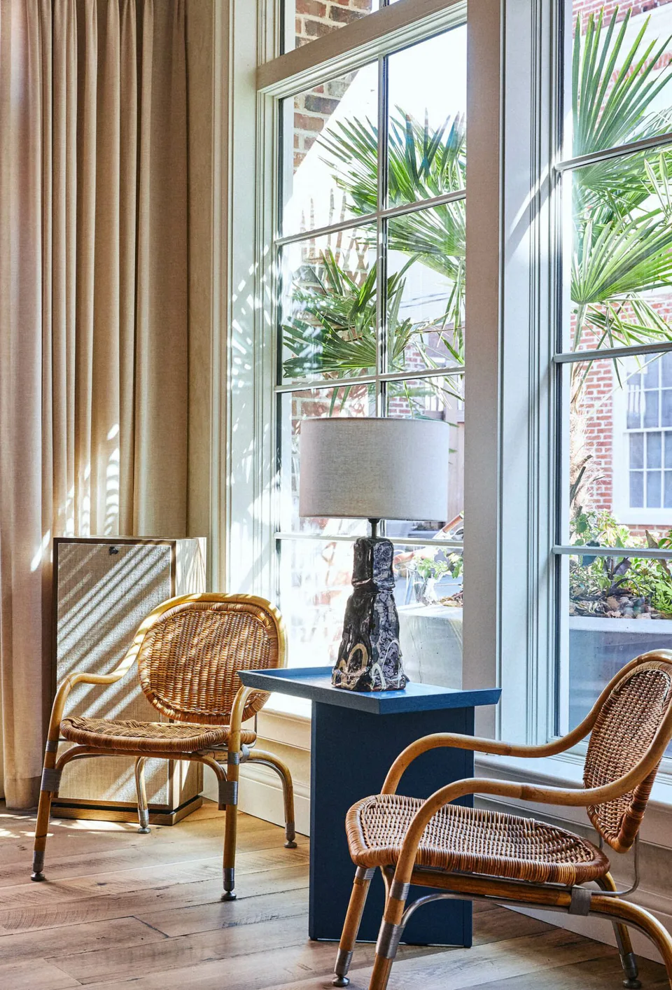Rattan armchairs and a blue side table with a sculptural lamp by the tall lobby windows at The Pinch Hotel, Charleston, South Carolina, interior design by Alexandra Peck Design