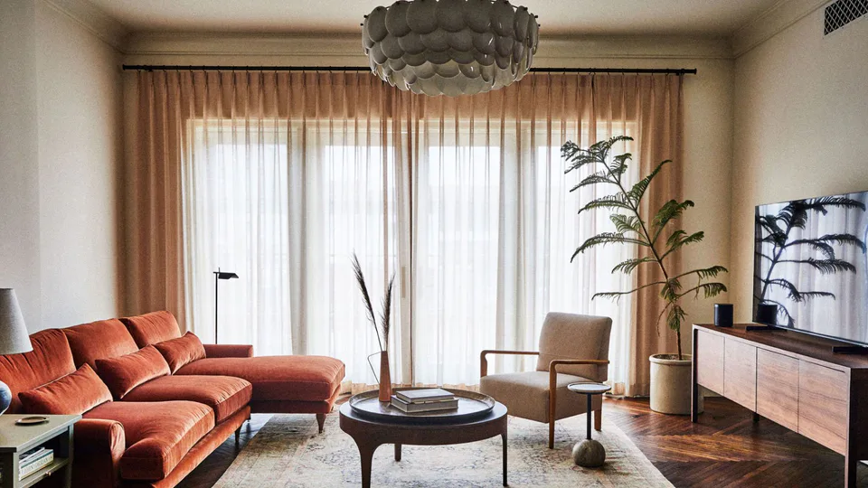 Suite living room with a rust velvet sectional, a round coffee table, a sculptural pendant and full-height curtains at The Pinch Hotel, Charleston, South Carolina, interior design by Alexandra Peck Design
